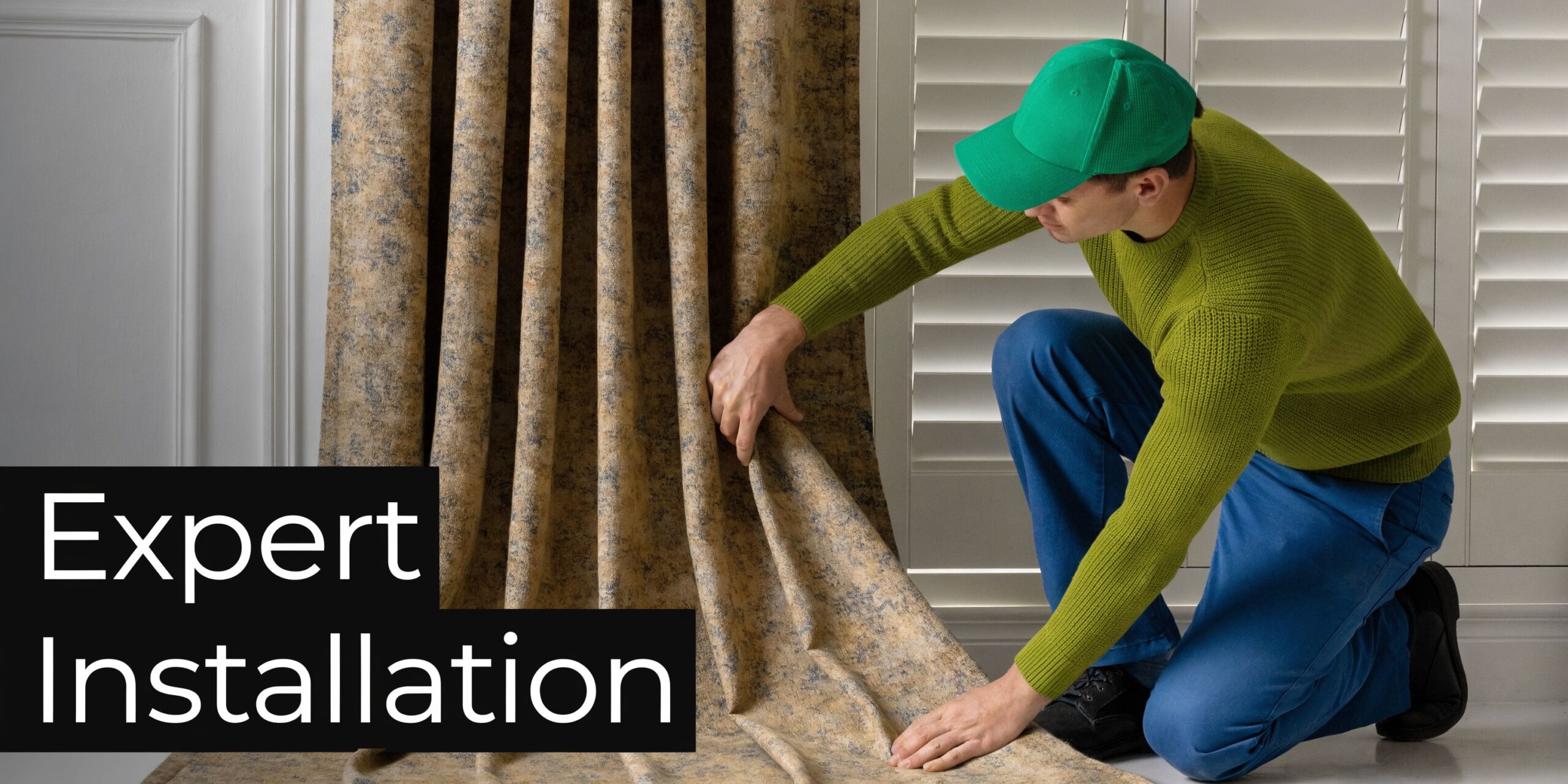 A professional installer in a green cap and sweater carefully adjusting long drapes next to white shutters.